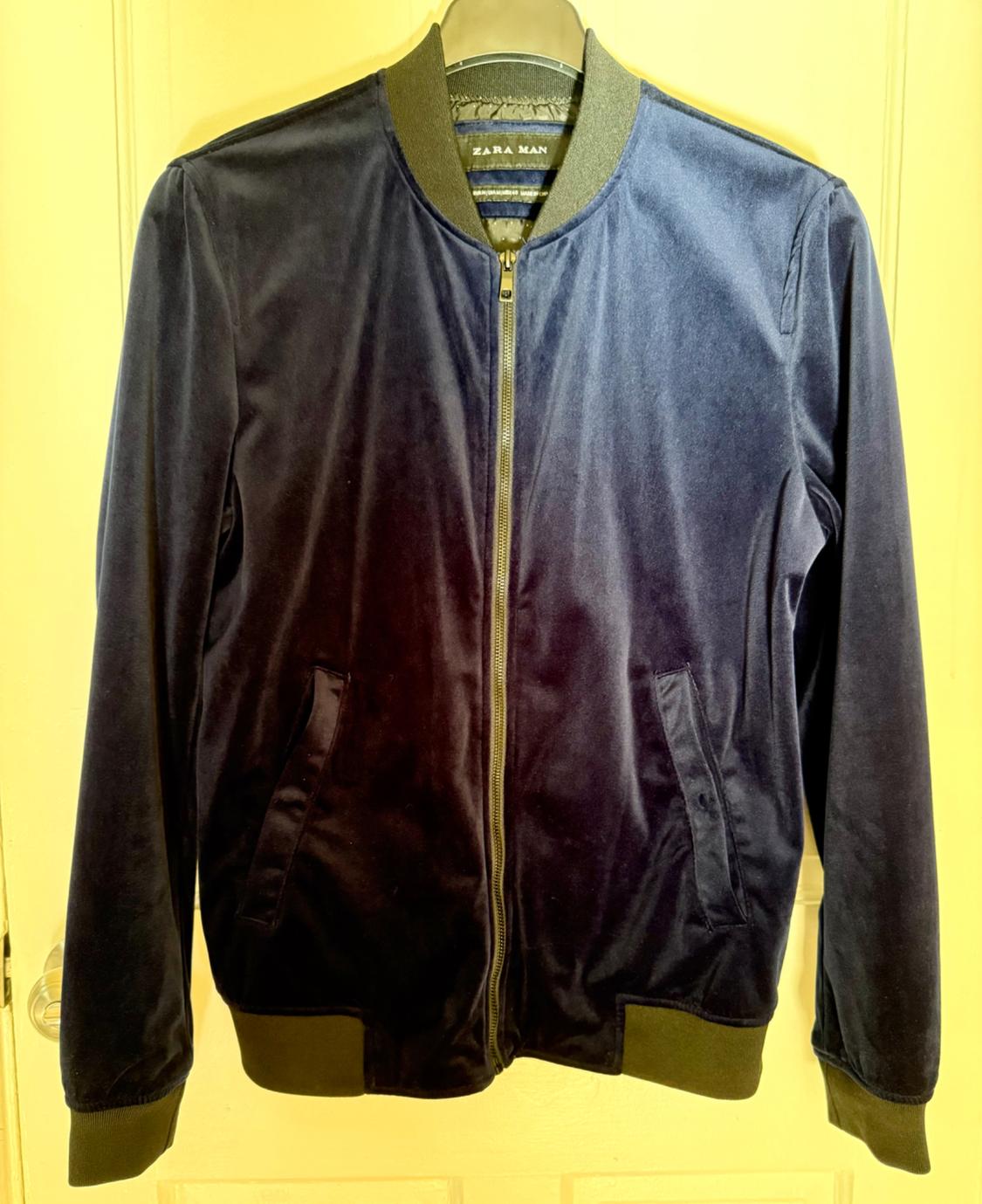 Navy blue velvet bomber jacket with a zipper and elastic collar, hanging on a door. Interior label with the text ZARA MAN.