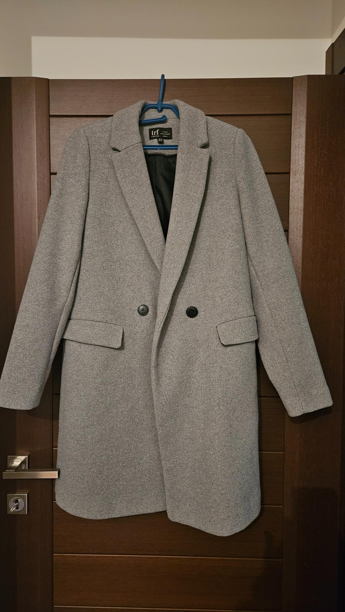 Grey wool coat hanging on a wooden door. It has wide lapels, two black buttons, and flap pockets. Label "trf".