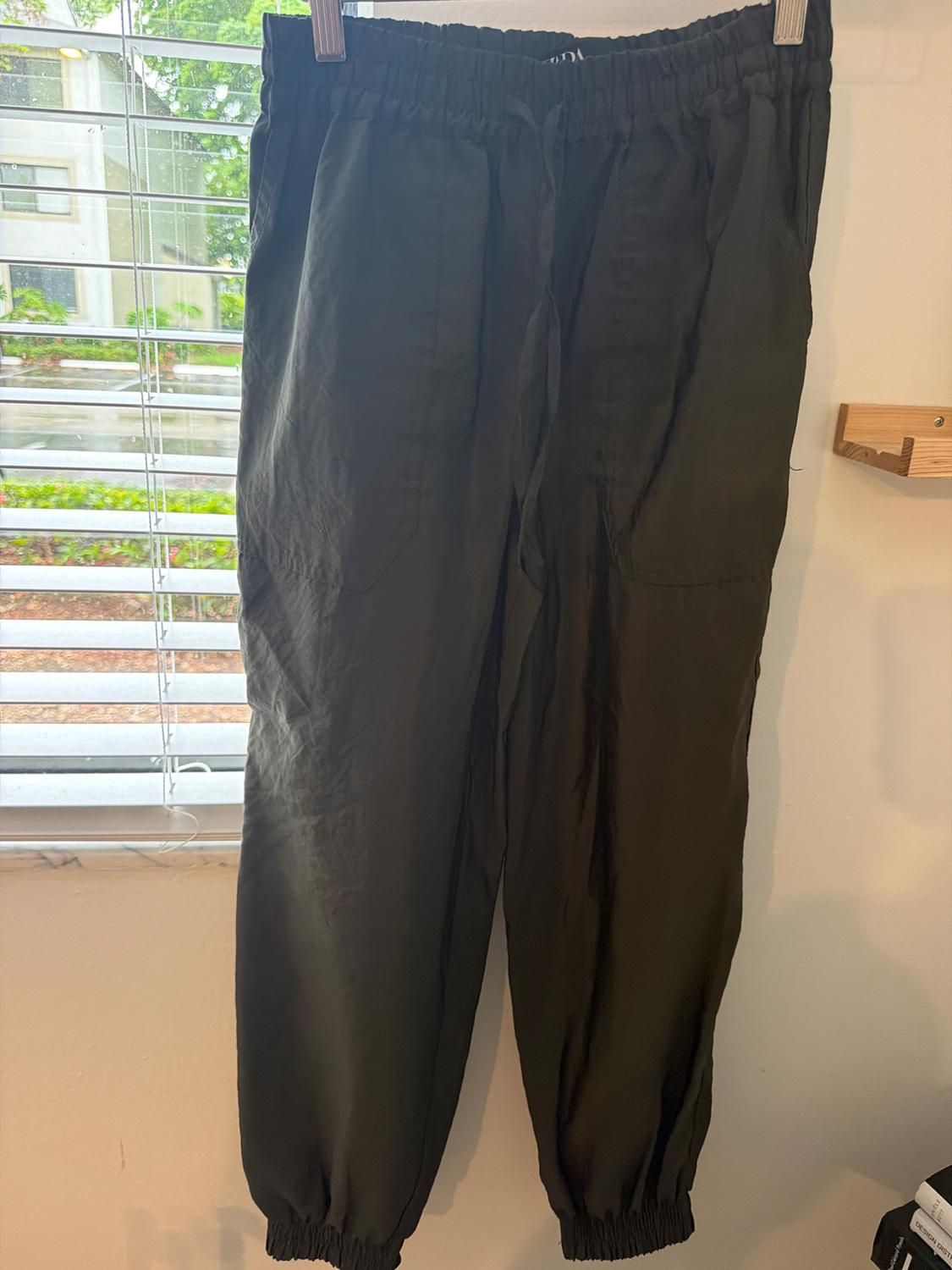 Dark green joggers with an elastic waistband and cuffs, hanging on a clip hanger next to a window with white blinds.