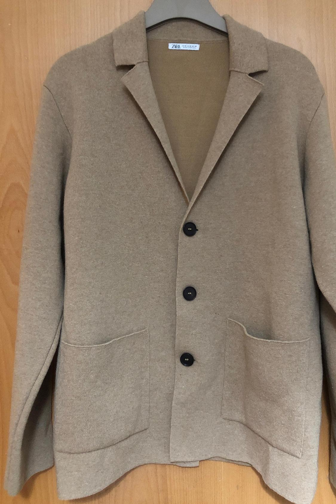 Front view of a beige jacket with lapels, black buttons and front pockets.