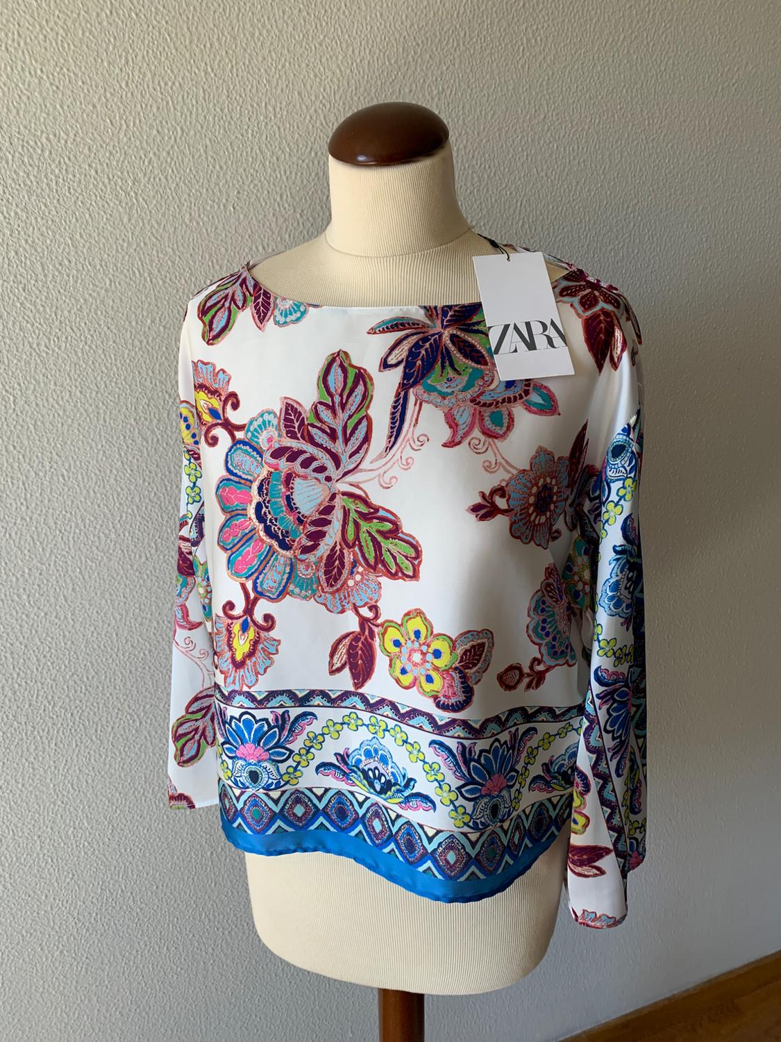 Long-sleeved white blouse with a colourful floral print and a blue border at the hem, displayed on a mannequin. Label with the text ZARA.