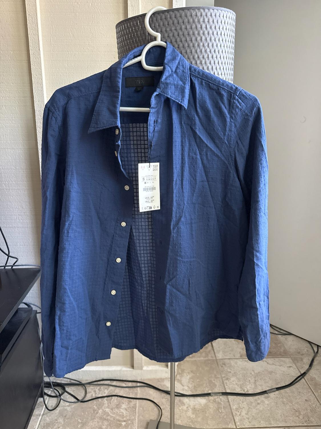 Blue long-sleeved shirt hanging on a white hanger. The black ZARA label is visible on the collar, along with a paper label showing size S.