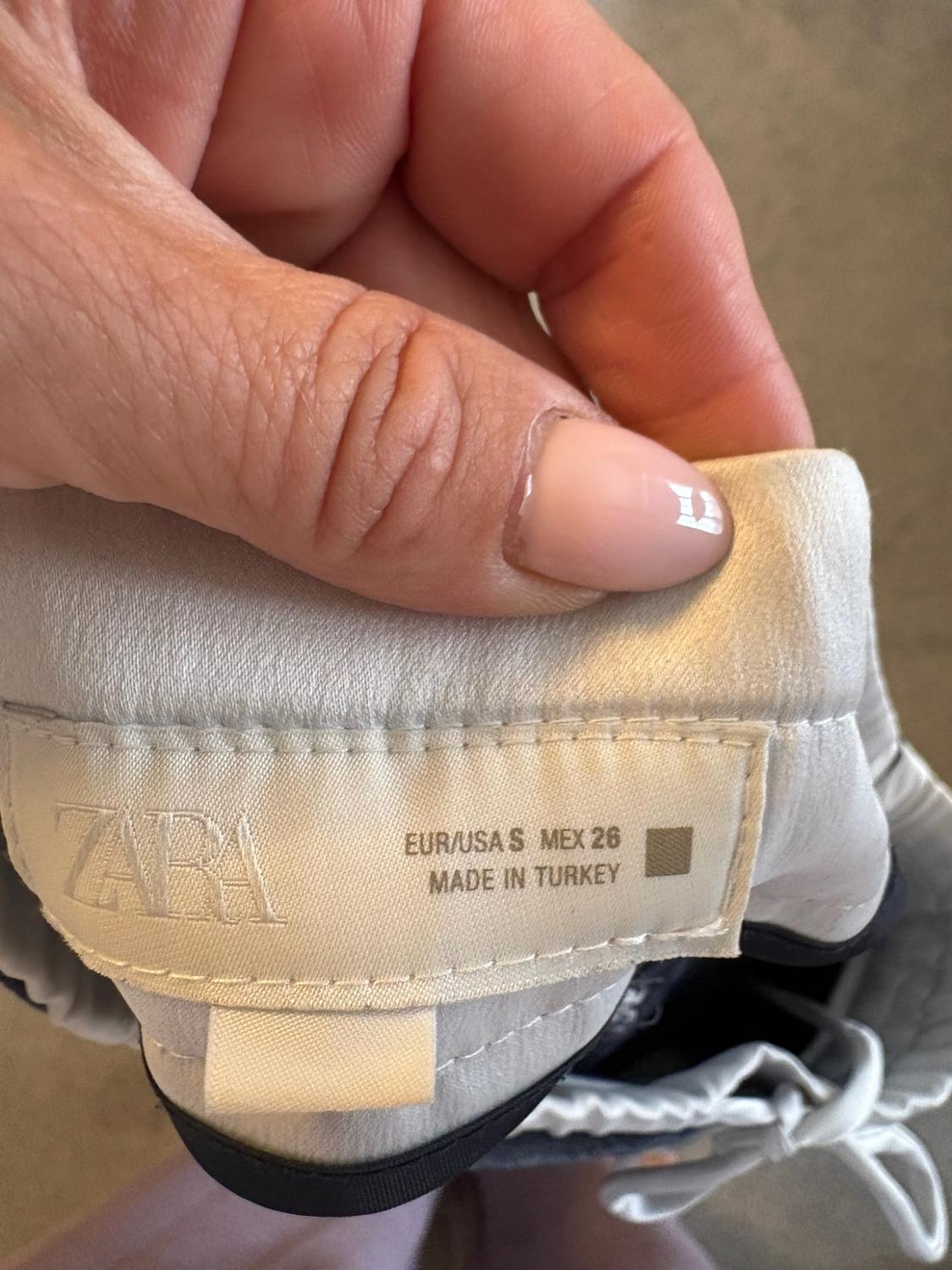 Here is the rephrased sentence, keeping the same meaning and context, without using any of the prohibited terms: "Close-up view of a hand holding a grey garment label. Text: ZARA EUR/USA S MEX 26 MADE IN TURKEY."