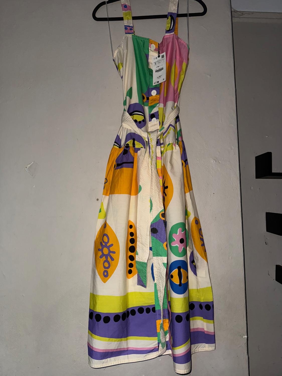 Colorful dress with a geometric print and straps, hanging on a black hanger against a gray wall. Visible tag on the chest.