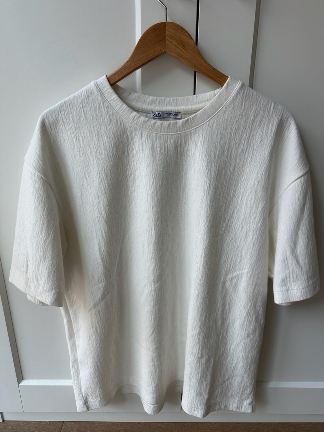 White short-sleeve t-shirt with texture, hanging on a wooden hanger in front of a white door. Label with the text ZARA.