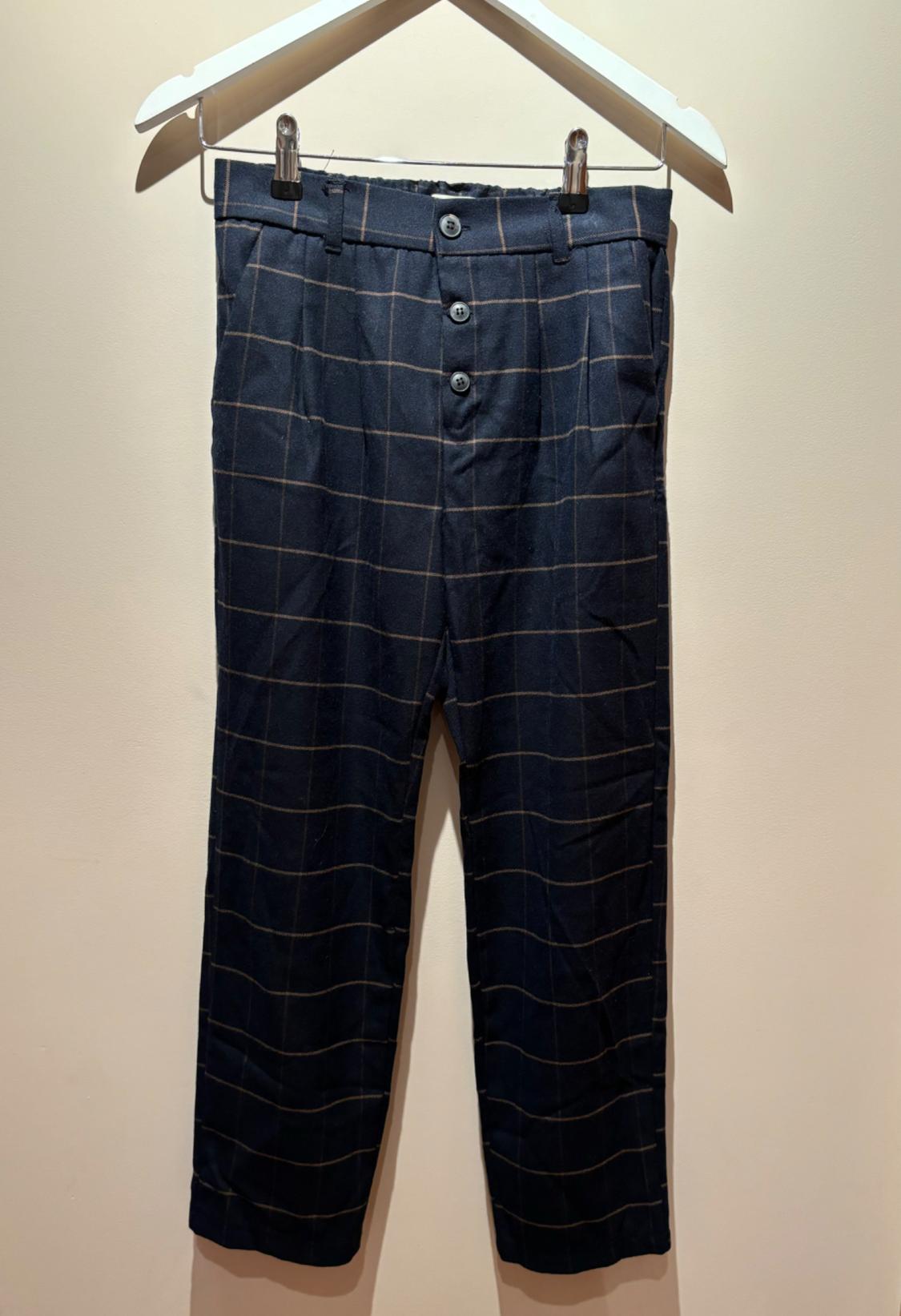 Dark blue trousers with a fine check pattern in beige lines, three buttons at the front, hanging on a white hanger against a beige background.
