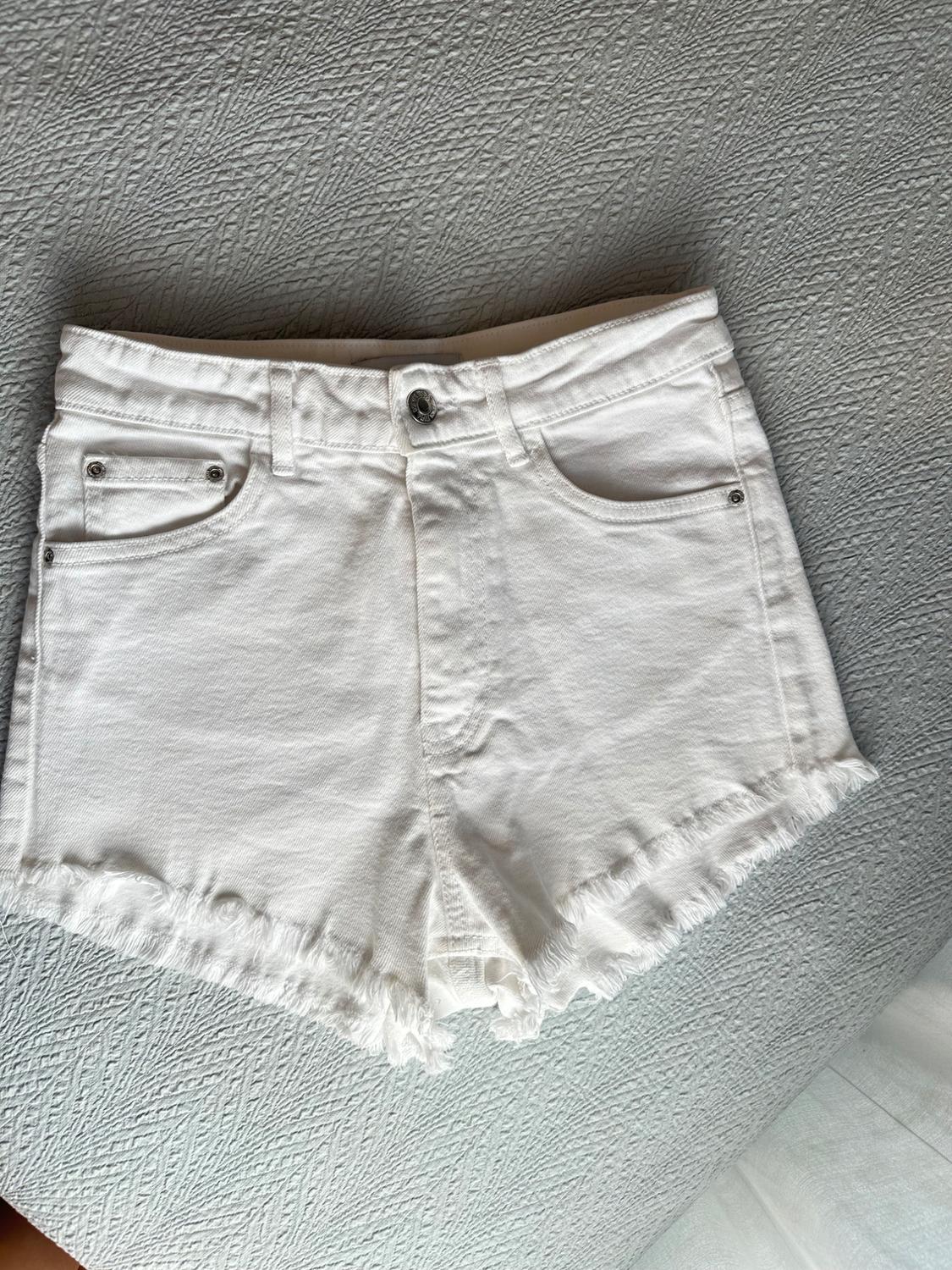White denim shorts with pockets and frayed hem, on a textured grey fabric surface. Casual style.