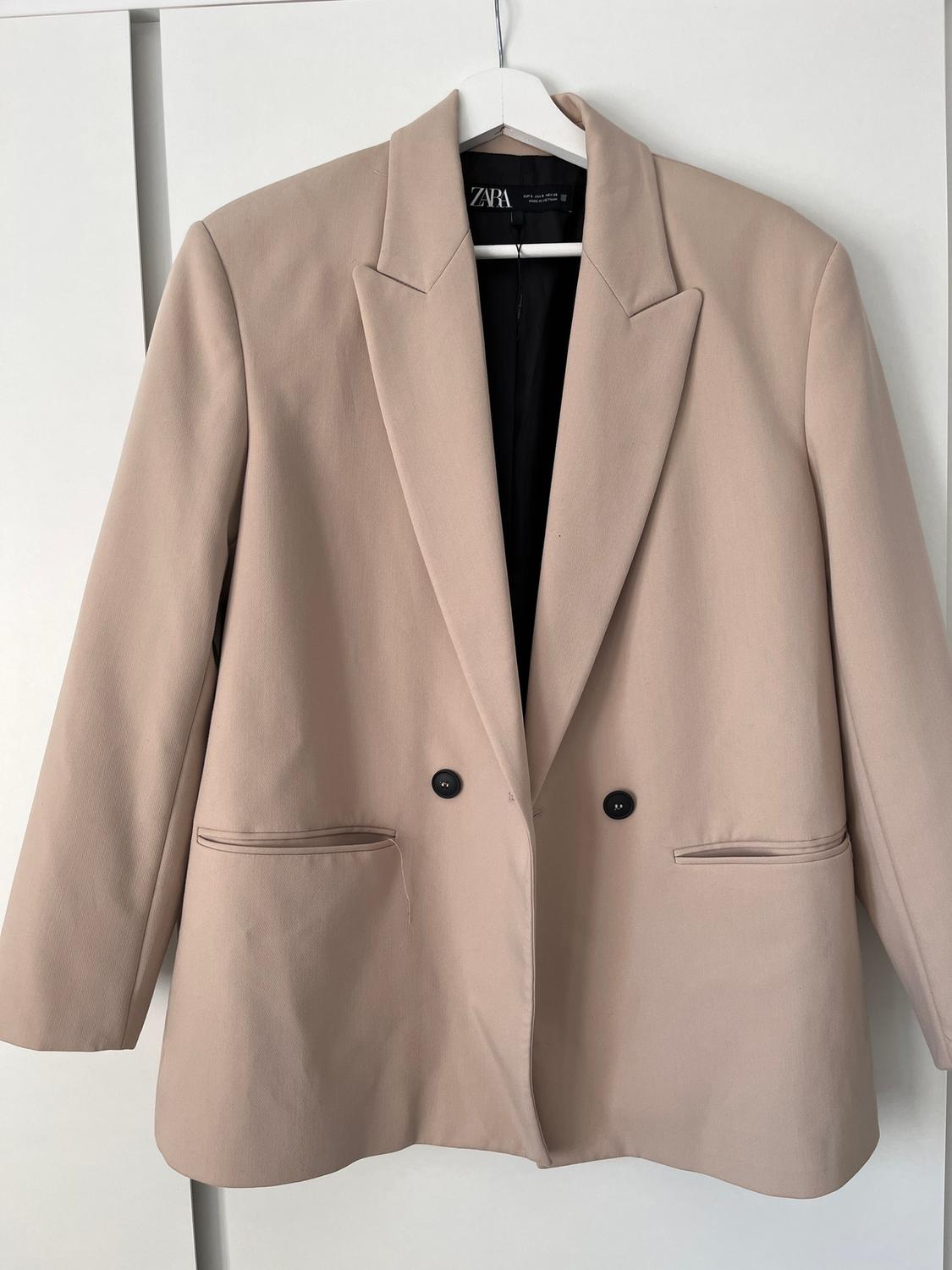 Beige double-breasted blazer hanging on a white hanger. Visible "ZARA" brand label on the inside of the collar.
