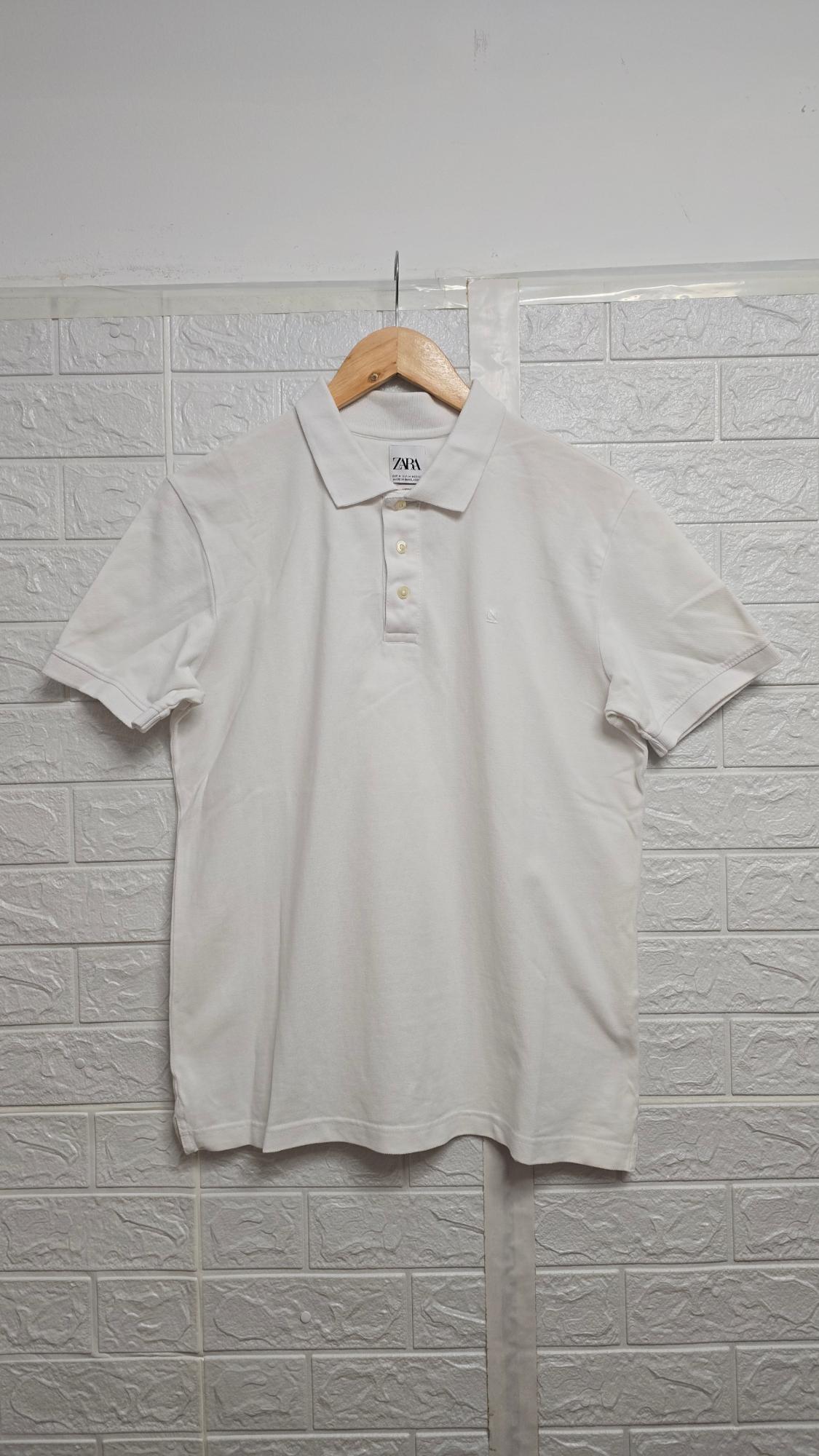 White polo shirt hanging on a wooden coat hanger in front of a white brick wall. Label visible with the text "ZARA".