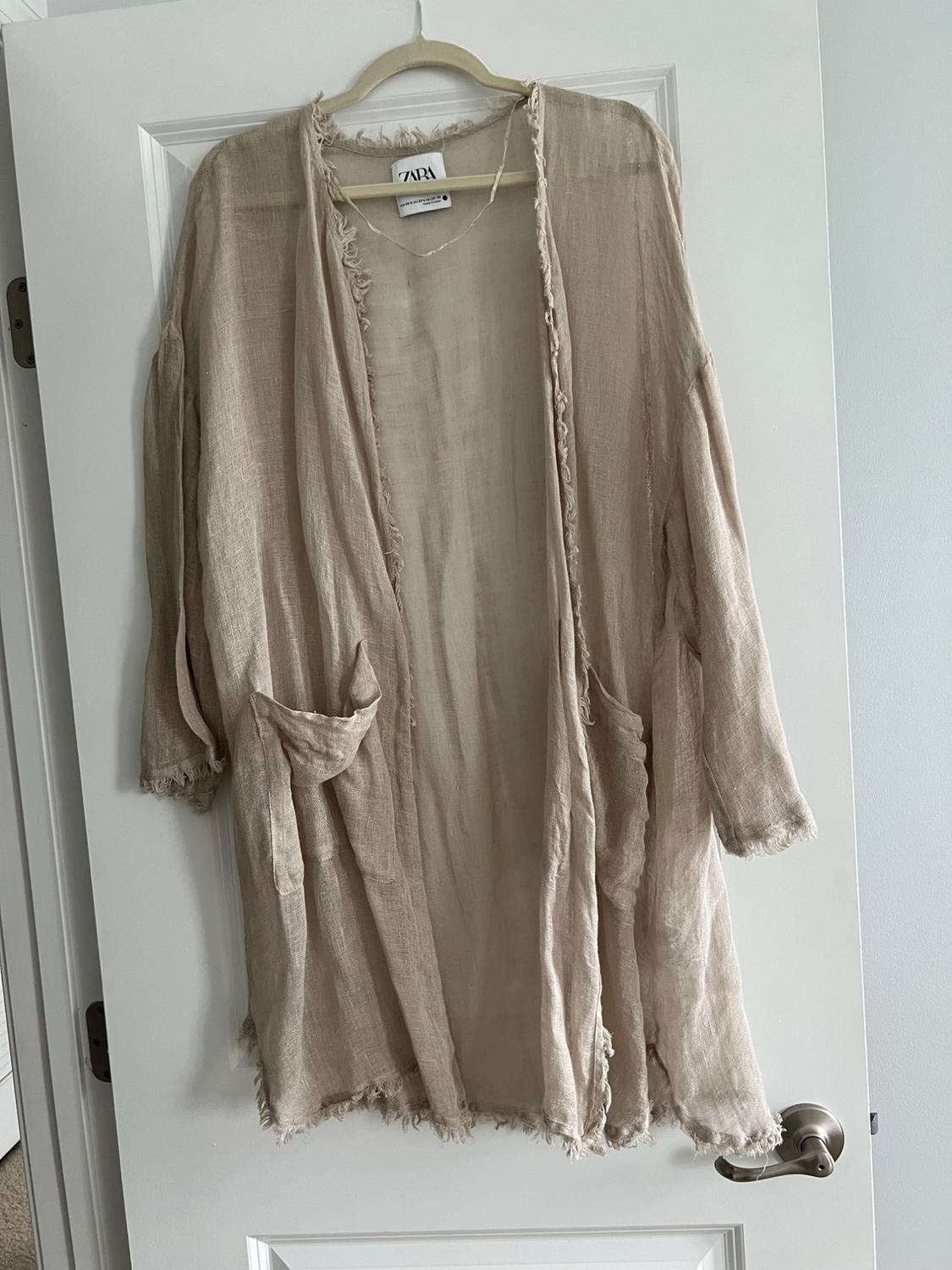 Beige cardigan made of lightweight fabric with frayed edges, hanging on a white door. Visible tag with the text "ZARA".