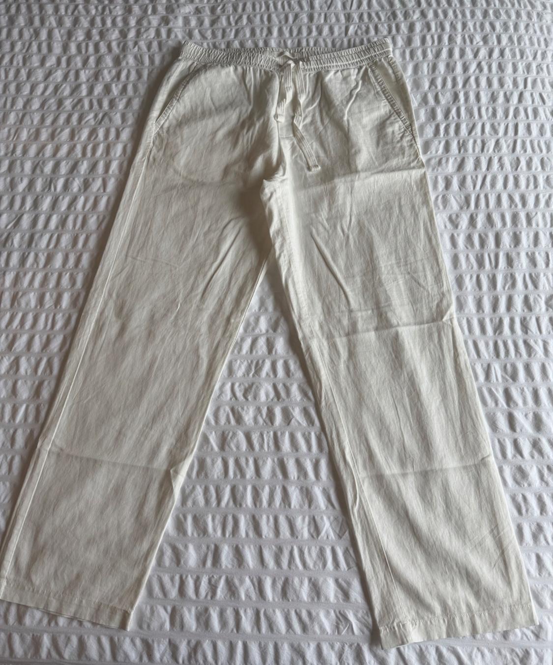 Casual white pants with an elastic waistband and drawstring, laid out on a textured white bedspread. They convey a relaxed style.