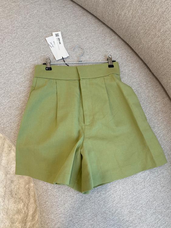 Olive green shorts on a beige sofa, hanging on a hanger with visible tags.