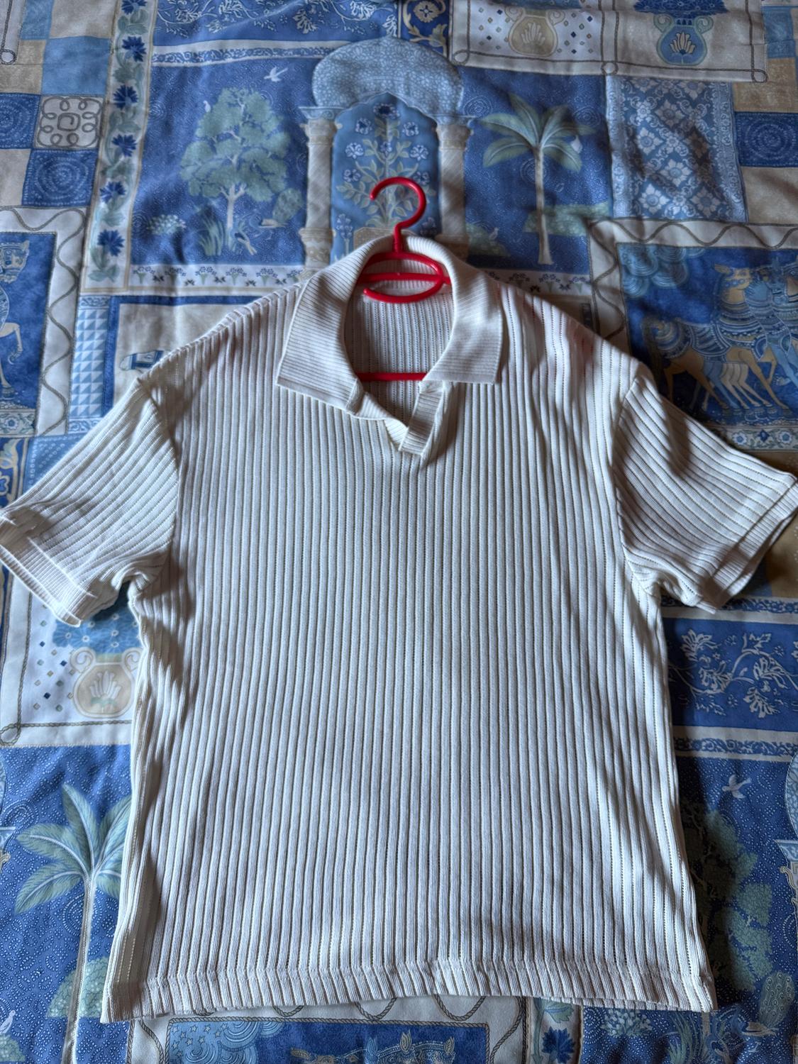 Beige knitted polo shirt with a ribbed texture, hanging on a red hanger over a blue and beige patterned bedspread.
