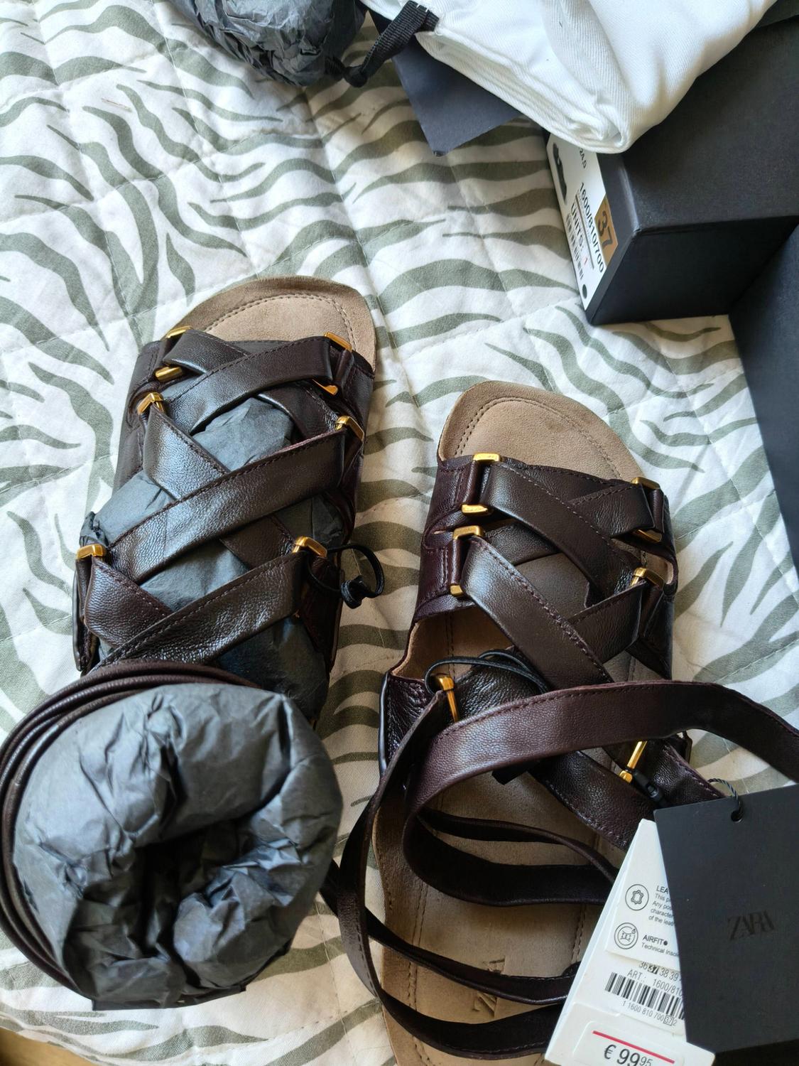 Brown strappy sandals on zebra-print fabric. A box and labels bearing the text ZARA, AIRFIT, 37, 1600/810, €99.95 can be seen.