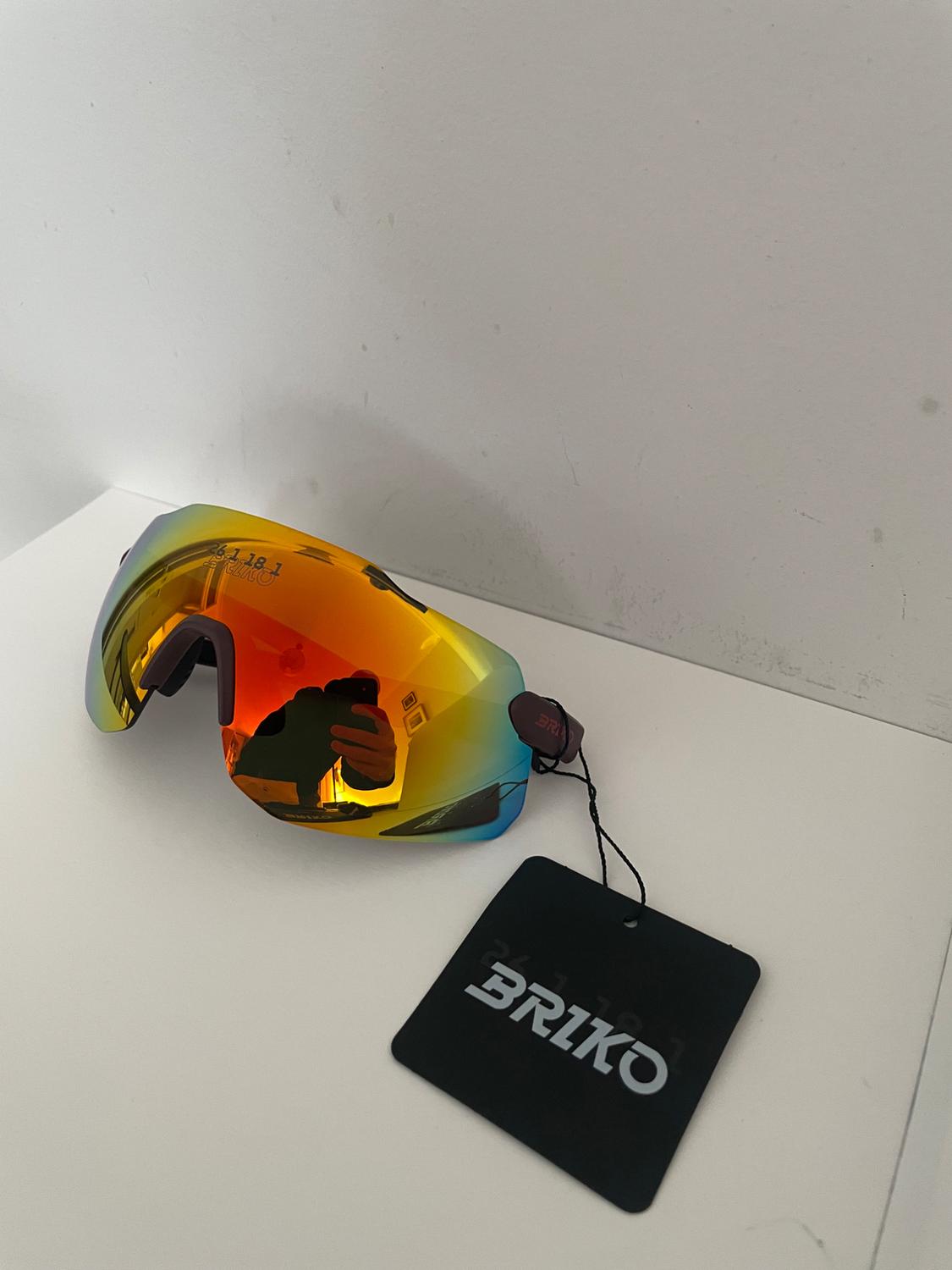 Sports glasses with an orange mirrored lens on a white background. Black label with the text BRIKO. Text on lens: 26.1.18.1 BRIKO.