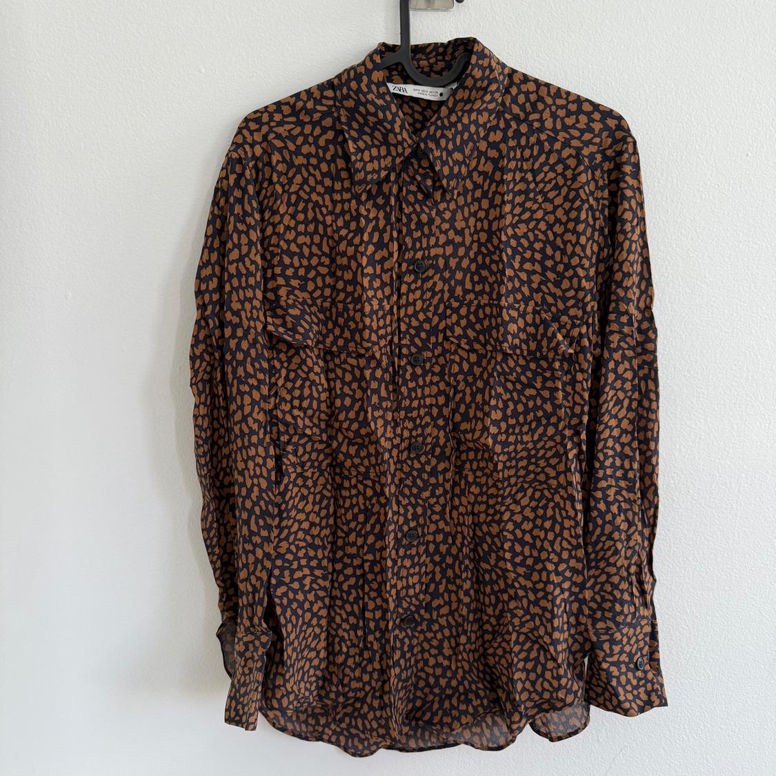 Long-sleeved shirt with a brown spot print on a black background, hanging on a hanger against a white wall.