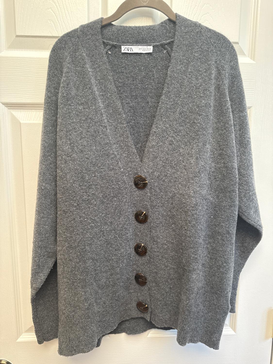 Grey knitted cardigan with dark buttons, hanging on a hanger in front of a white door. Visible label: Zara.