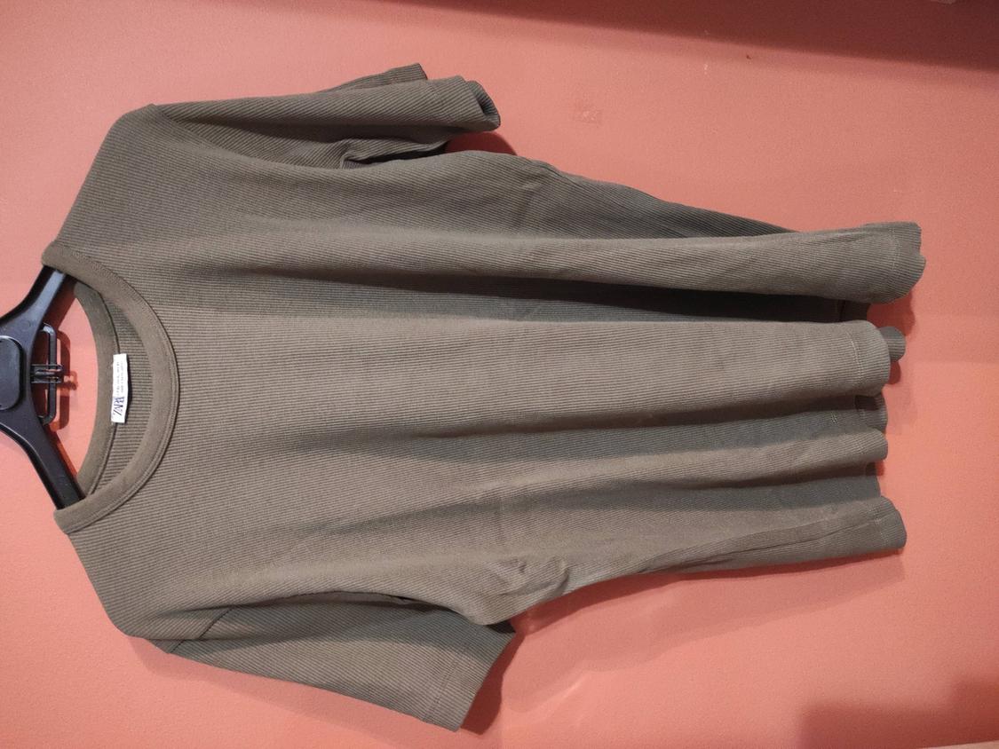 Khaki short-sleeved T-shirt hanging on a black hanger against a pink wall. Label visible on the collar.