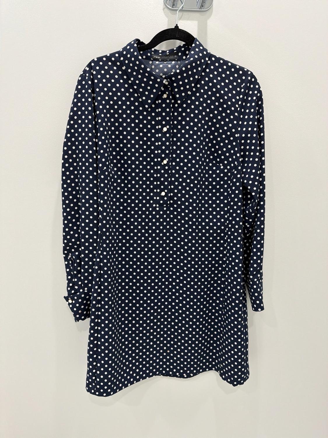 Navy blue dress with white polka dots, collar and buttons, hanging on a black hanger against a white background. Text: ZARA.