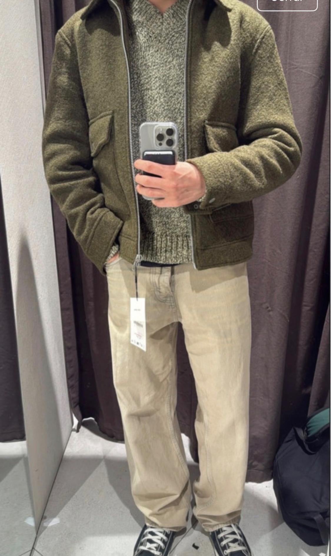 Fitting room selfie. Person wearing an olive green jacket, marl knit jumper, beige jeans with a tag, and black trainers.