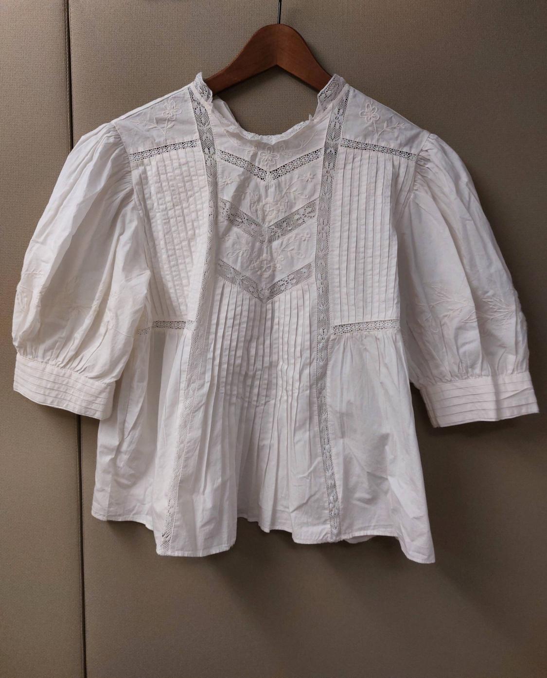 White blouse with puffed sleeves, lace detailing and embroidery, hanging on a wooden hanger against a neutral background.