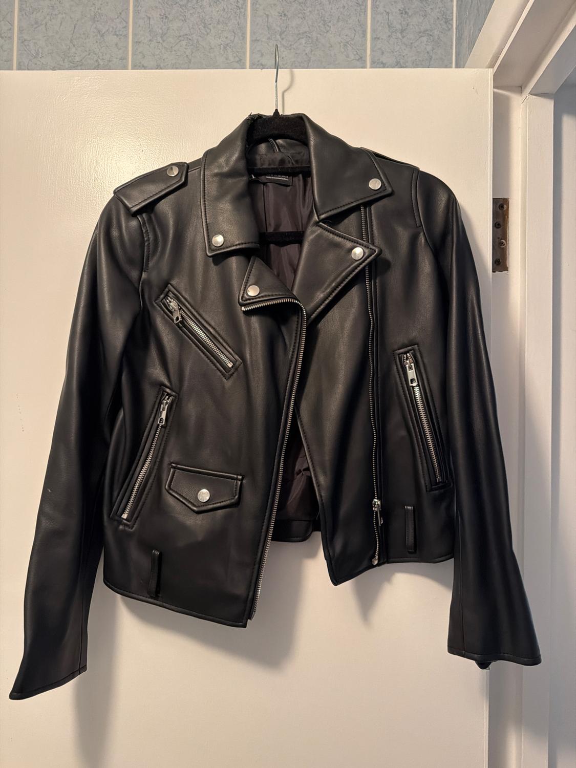 Black leather jacket with zips and silver buttons, hanging on a white door. Indoor setting with soft lighting.