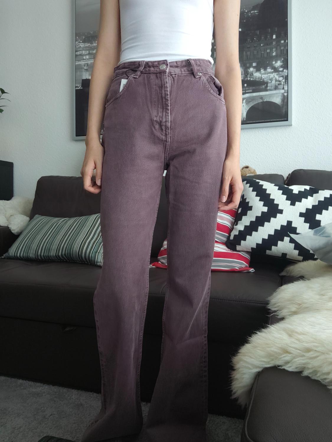 Person in a living room wearing a white top and high-waisted straight-leg purple jeans. In the background, a dark sofa with cushions.