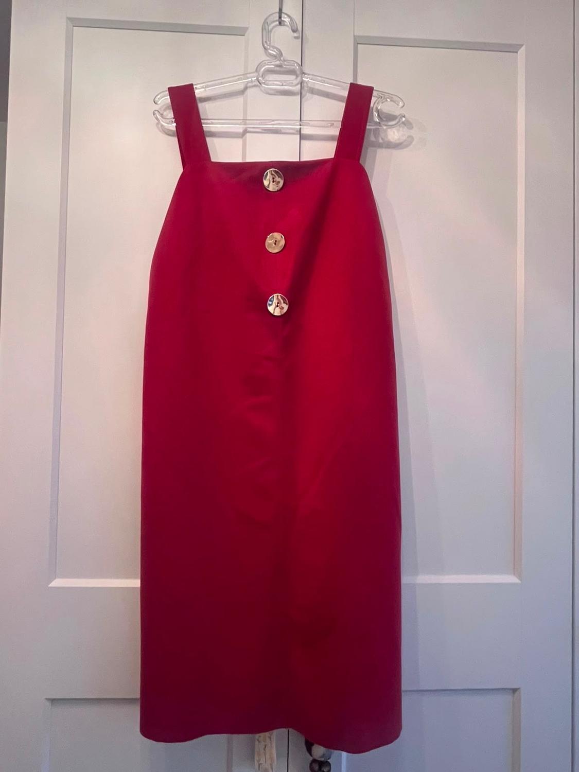 Red dress with wide straps and three front buttons, hanging on a clear hanger over a white door.