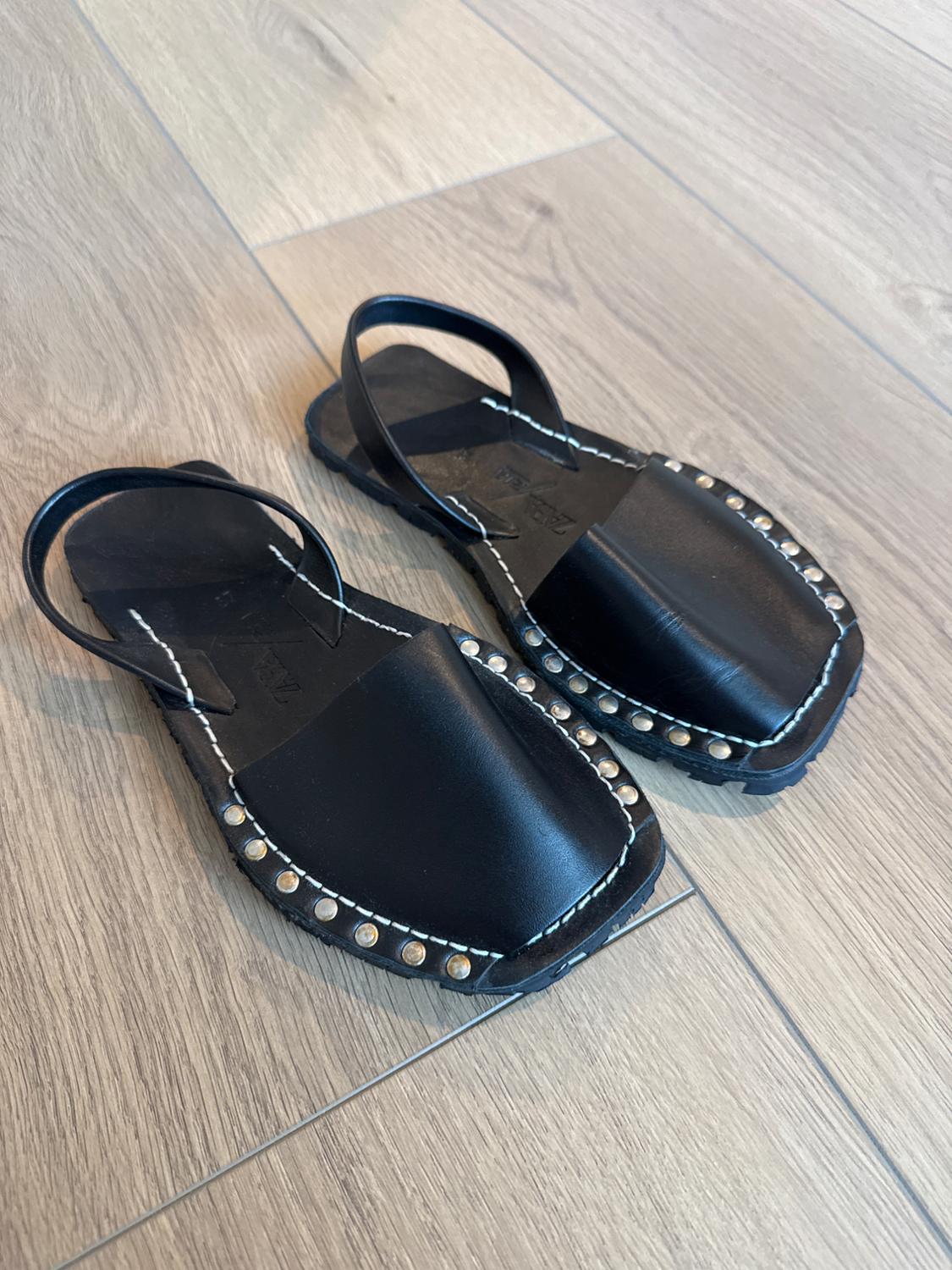 Black sandals with studs, white stitching, and a back strap on a wooden floor. ZARA text on the insole.