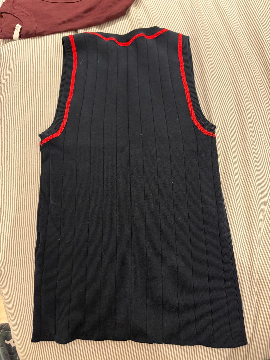 Black sleeveless waistcoat with red trim on a striped fabric surface. Part of a burgundy garment visible in the corner.