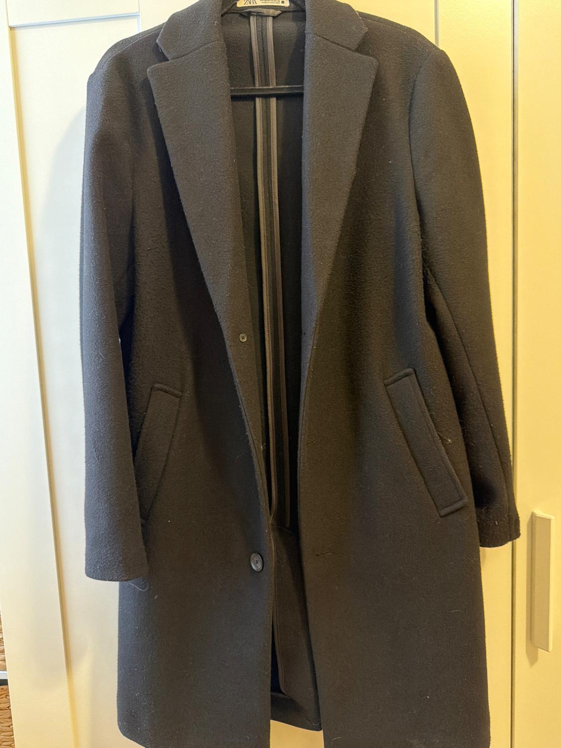 Black coat hanging on a hanger in front of a white door. Features a lapel collar, buttons and side pockets. ZARA label on the inside.