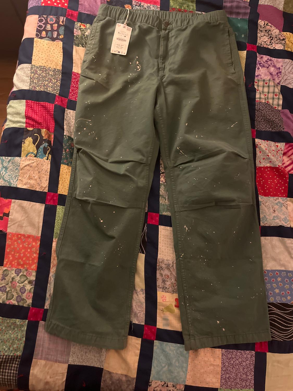 Green trousers with white paint splatters on a colourful patchwork quilt. Hang tag visible at the waist.