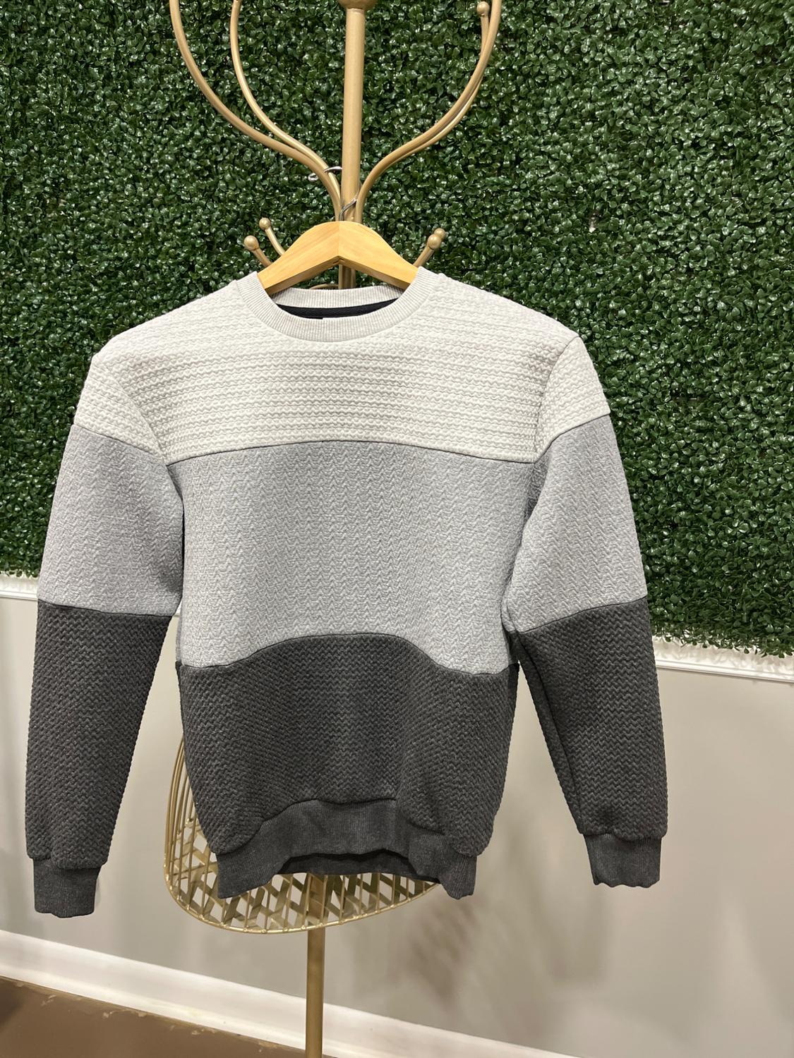 Knit sweater in gray tones hanging on a golden rack, with a green artificial leaf wall background.