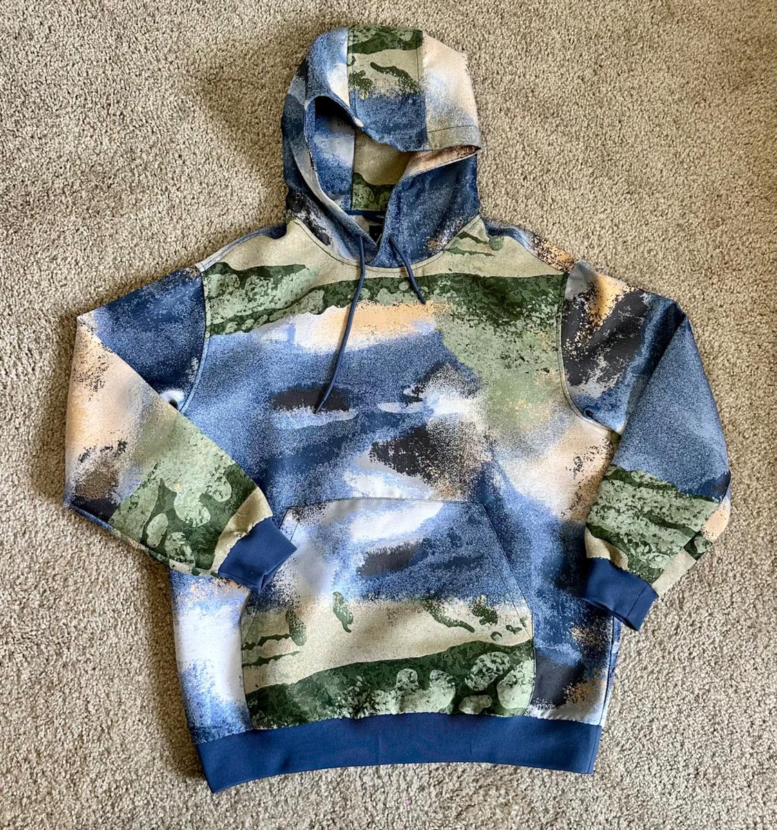 Casual hoodie, blue, green and beige abstract print, front pocket, blue cuffs and hem, on a beige rug.