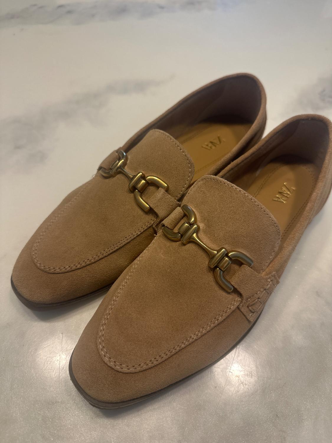 Light brown suede shoes with gold metallic detailing on the upper, on a white marble surface.