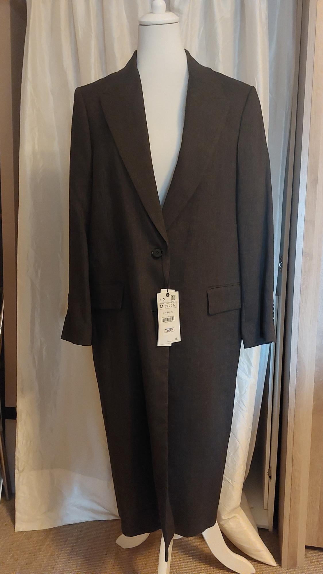 Dark brown coat on a white mannequin, with visible label indicating size M. White curtain background.