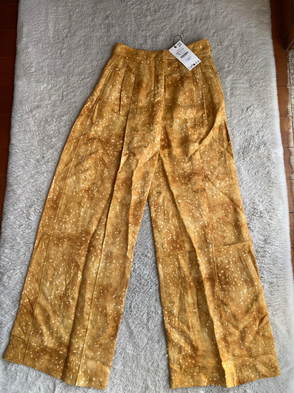 Mustard wide-leg trousers with a white spotted print, pleats and a label, laid out on a soft-textured white carpet.