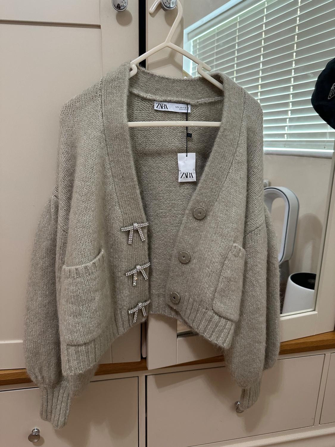 Chunky knit beige cardigan with large buttons and shiny decorative bows, hanging on a hanger in front of a wardrobe.