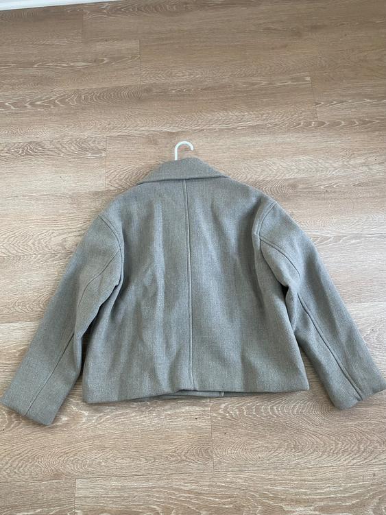 Gray thick fabric jacket seen from behind, hanging on a white hanger over a light wood floor.