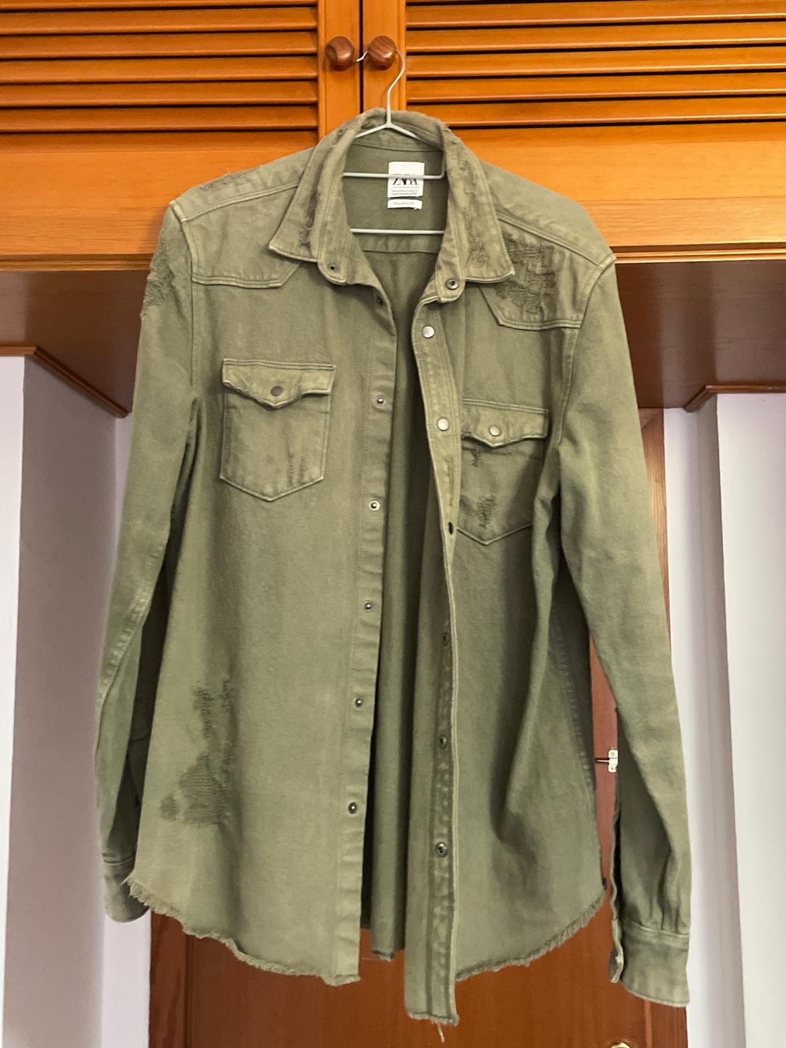 Olive green long-sleeved shirt with chest pockets, hanging on a hanger in front of a wooden door.
