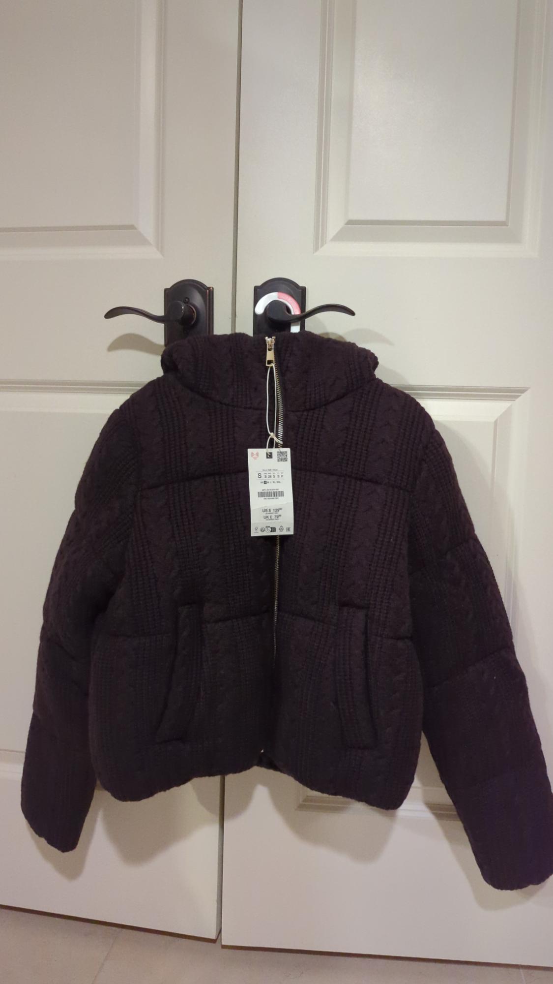 Dark brown padded jacket with a hood and zip, hanging on a white door. Visible label with product details.