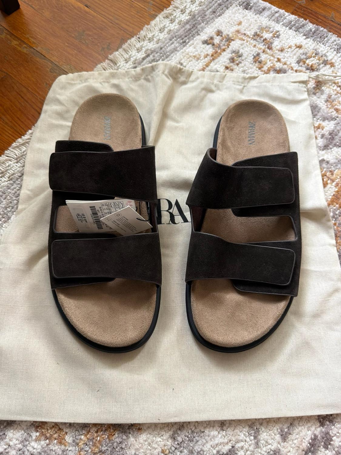 Brown two-strap sandals with tag and ZARA MAN text on the insole,
resting on a fabric bag and a textured rug.