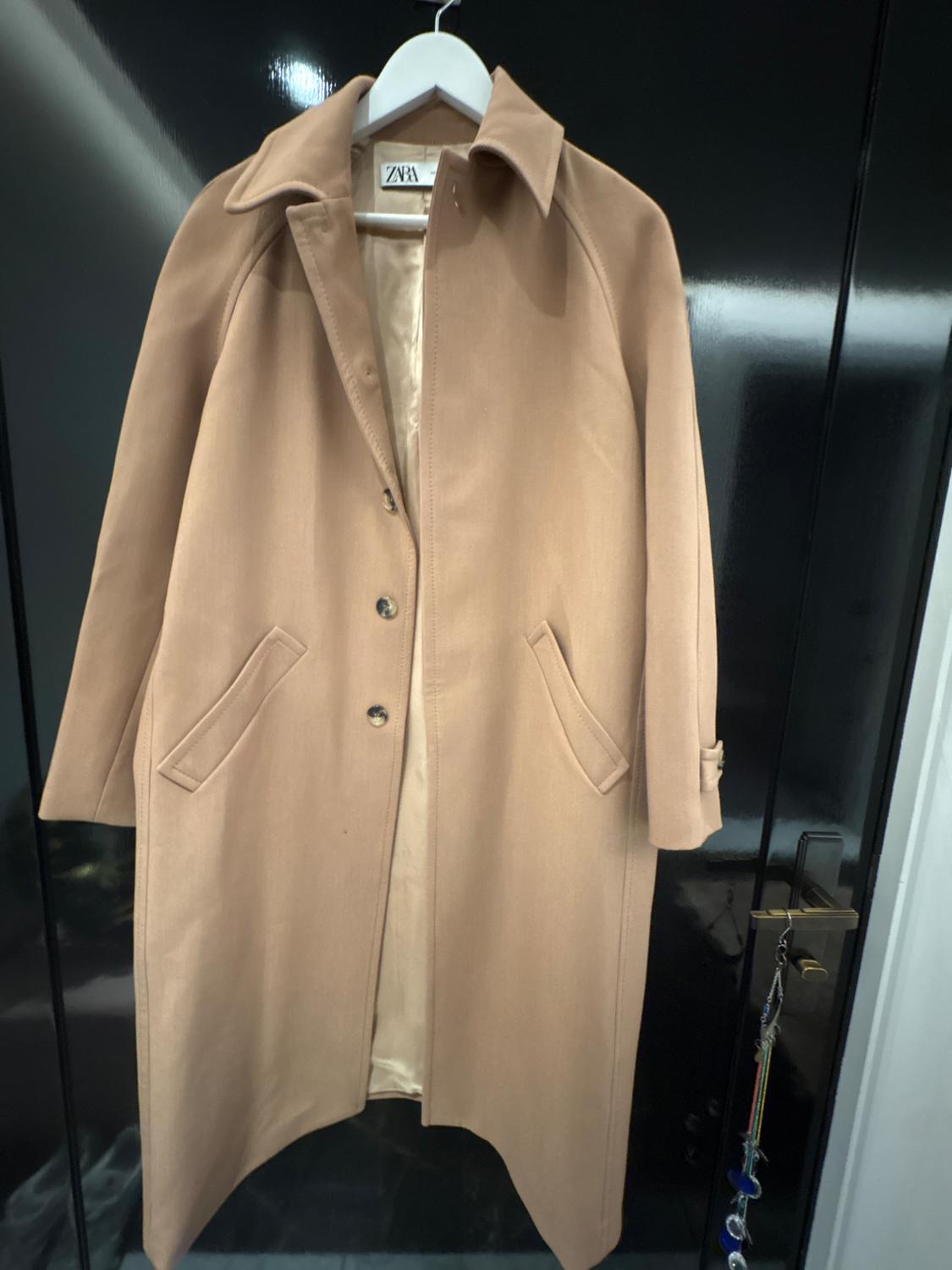 Beige coat hanging on a white hanger, with a visible "ZARA" tag, against a dark background and a door with a keyring.