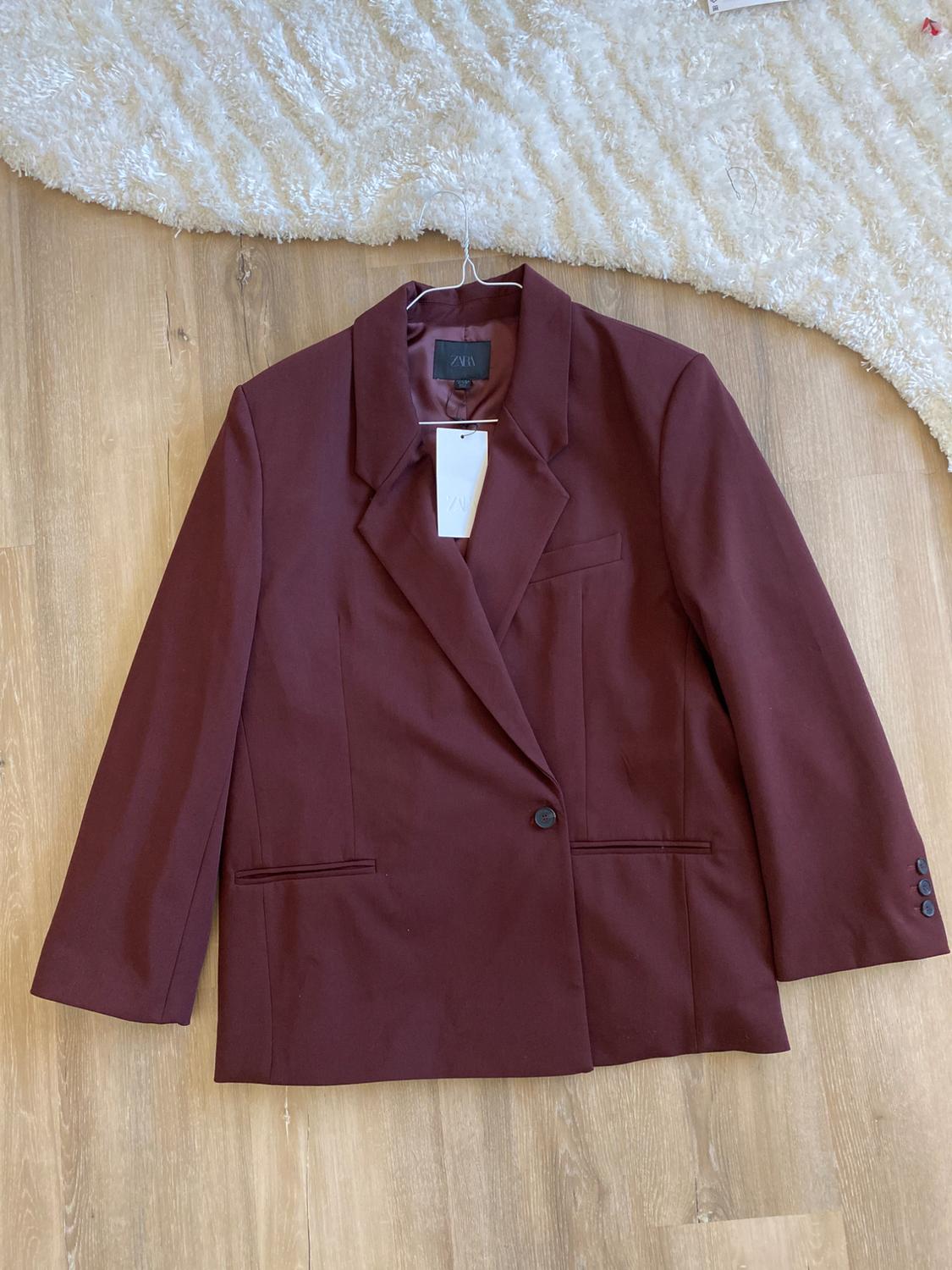 Single-button brown jacket hanging on a hanger over a wooden floor and white rug. Label visible on the collar.