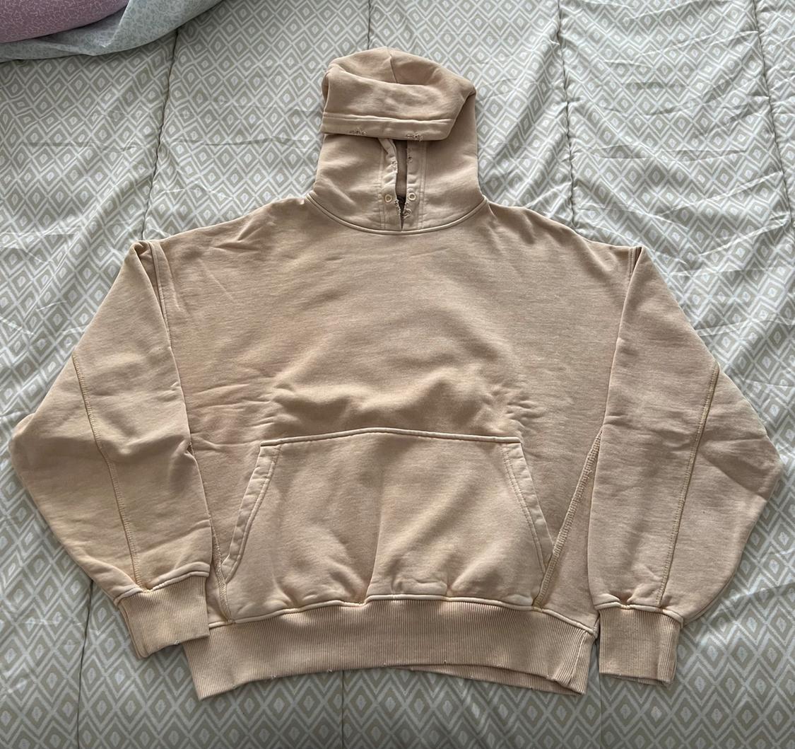 Beige hoodie with a front pocket, placed on a quilt with a diamond pattern in light tones.
