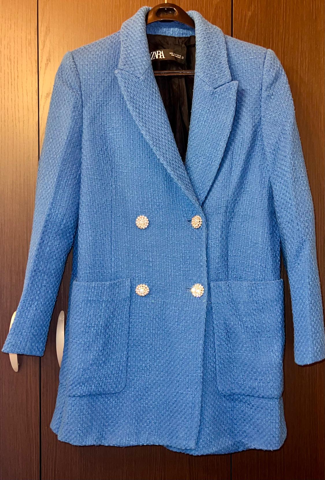 Textured blue coat with shiny decorative buttons and large pockets, hanging on a hanger in front of a wooden background.