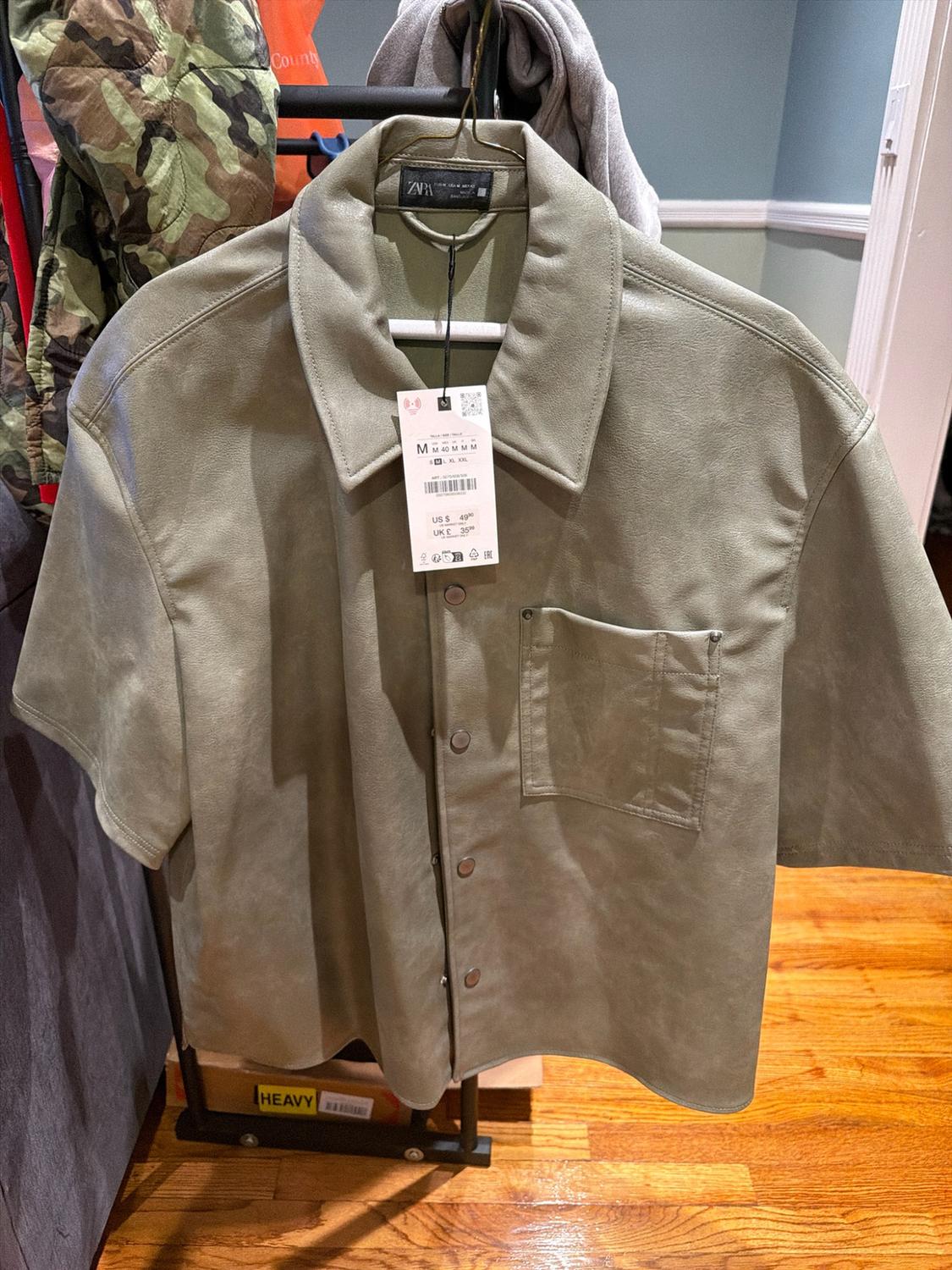 Olive green short-sleeve Zara shirt, with a collar, buttons, a chest pocket, and a price tag, hanging on a hanger.