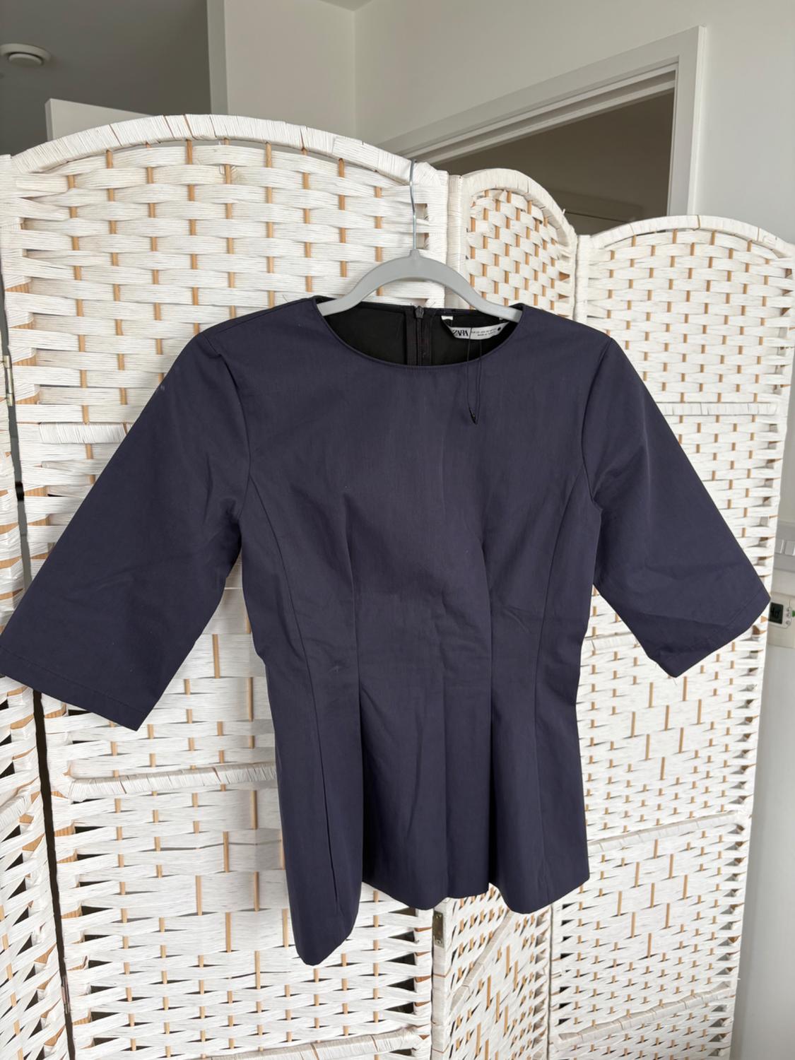 A dark blue, three-quarter sleeve blouse is hanging on a white coat rack, in front of a white wicker screen in an interior.