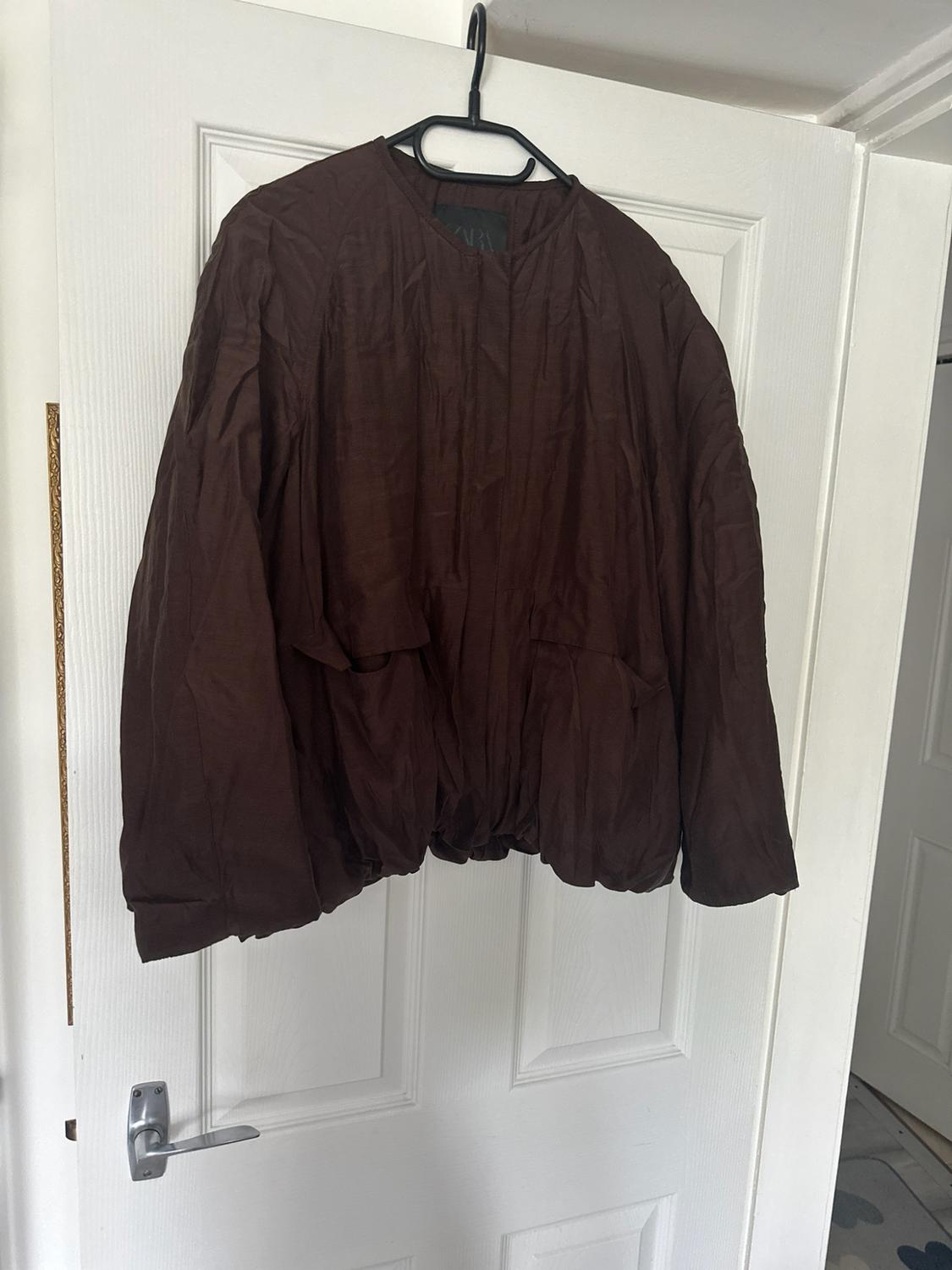 Dark brown long-sleeved jacket hanging on a hanger over a white interior door. ZARA label on the collar.