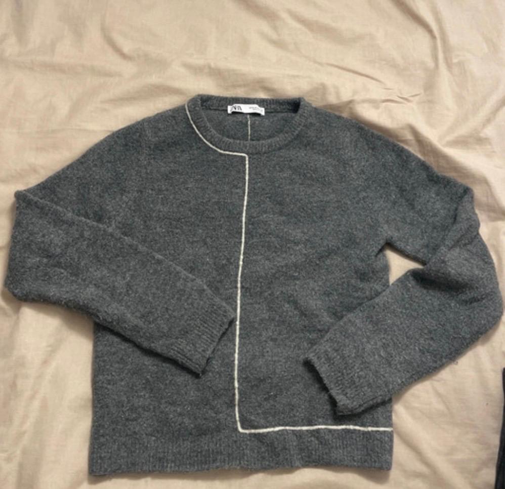 Grey jumper with a white line in the middle, on a beige surface. Visible label on the collar.